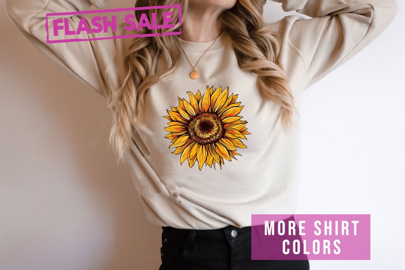 May include: A beige sweatshirt with a yellow sunflower graphic. The text "FLASH SALE" is in pink at the top of the image. The text "MORE SHIRT COLORS" is in pink at the bottom of the image.