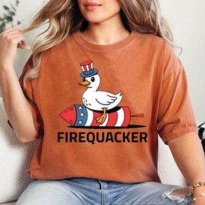 Peut inclure: T-shirt orange br&ucirc;l&eacute; avec un canard de dessin anim&eacute; portant un chapeau patriotique, chevauchant un feu d'artifice rouge, blanc et bleu. Le texte "FIREQUACKER" est imprim&eacute; en noir en dessous. Le t-shirt est en mati&egrave;re souple.