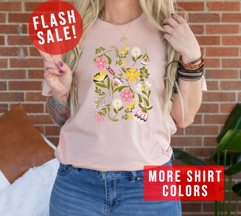 May include: A pink t-shirt with a floral print featuring pink, yellow, and white flowers. The shirt has a crew neck and short sleeves. The text "MORE SHIRT COLORS" is visible in the bottom right corner of the image.