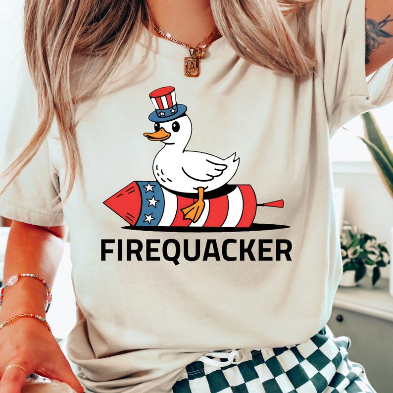 Peut inclure: T-shirt beige avec un motif d'un canard blanc portant un chapeau patriotique, assis sur une fus&eacute;e rouge, blanche et bleue. Le texte "FIREQUACKER" est imprim&eacute; en noir en dessous. Le motif est centr&eacute; sur le t-shirt.