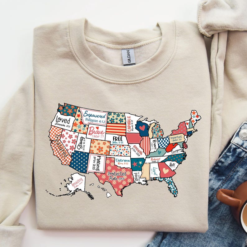 May include: A beige sweatshirt with a patchwork design of the United States map. Each state is filled with a different pattern and includes a word or phrase like "Loved", "Empowered", "Brave", "Free", "Capable", "Enough", "Protected", "Amazing", "Strong", "Precious", and "Loyal".
