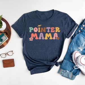 May include: A heather navy blue t-shirt with the text "POINTER MAMA" in colourful retro-style lettering. The shirt is laid out on a white surface with a pair of jeans, a pair of glasses, a watch, a camera, and a plant.