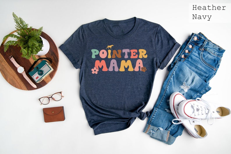 May include: A heather navy blue t-shirt with the text "POINTER MAMA" in colourful retro-style lettering. The shirt is laid out on a white surface with a pair of jeans, a pair of glasses, a watch, a camera, and a plant.