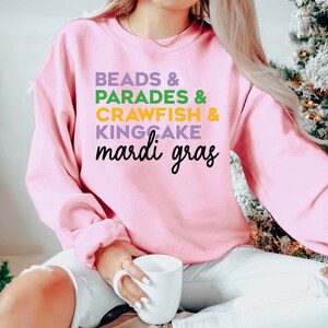 May include: Pink sweatshirt with the text "BEADS & PARADES & CRAWFISH & KINGCAKE mardi gras" in green, purple, and yellow lettering.
