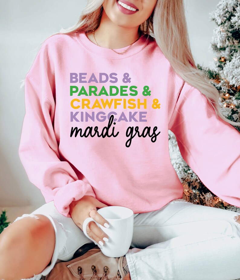 May include: Pink sweatshirt with the text "BEADS & PARADES & CRAWFISH & KINGCAKE mardi gras" in green, purple, and yellow lettering.