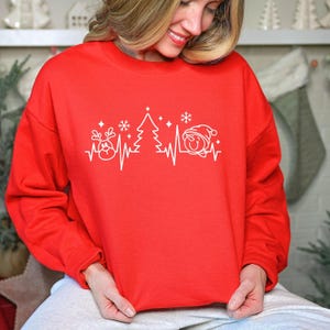 K&ouml;nnte beinhalten: Ein rotes Sweatshirt mit Rundhalsausschnitt und einem wei&szlig;en Grafikdesign mit Weihnachtselementen, darunter ein Rentier, ein Weihnachtsbaum und ein Schneemann, die entlang einer Herzschlaglinie angeordnet sind. Ein festliches Kleidungsst&uuml;ck.