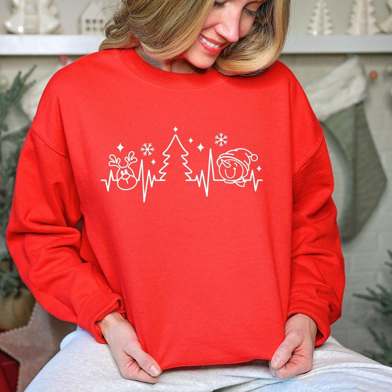 K&ouml;nnte beinhalten: Ein rotes Sweatshirt mit Rundhalsausschnitt und einem wei&szlig;en Grafikdesign mit Weihnachtselementen, darunter ein Rentier, ein Weihnachtsbaum und ein Schneemann, die entlang einer Herzschlaglinie angeordnet sind. Ein festliches Kleidungsst&uuml;ck.
