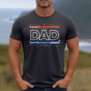May include: Dark gray t-shirt with the word "DAD" in white, stylized with a red lightsaber above and a blue lightsaber below. The design includes white speckles, creating a starry effect. The shirt is short-sleeved.