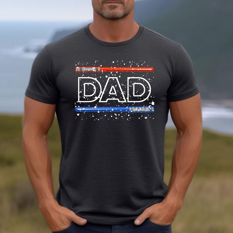 May include: Dark gray t-shirt with the word "DAD" in white, stylized with a red lightsaber above and a blue lightsaber below. The design includes white speckles, creating a starry effect. The shirt is short-sleeved.