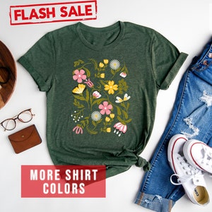 May include: Forest green t-shirt with a floral print featuring pink, yellow, and white flowers. The shirt is displayed with blue jeans and white sneakers. The text "FLASH SALE" and "MORE SHIRT COLORS" is visible in the image.