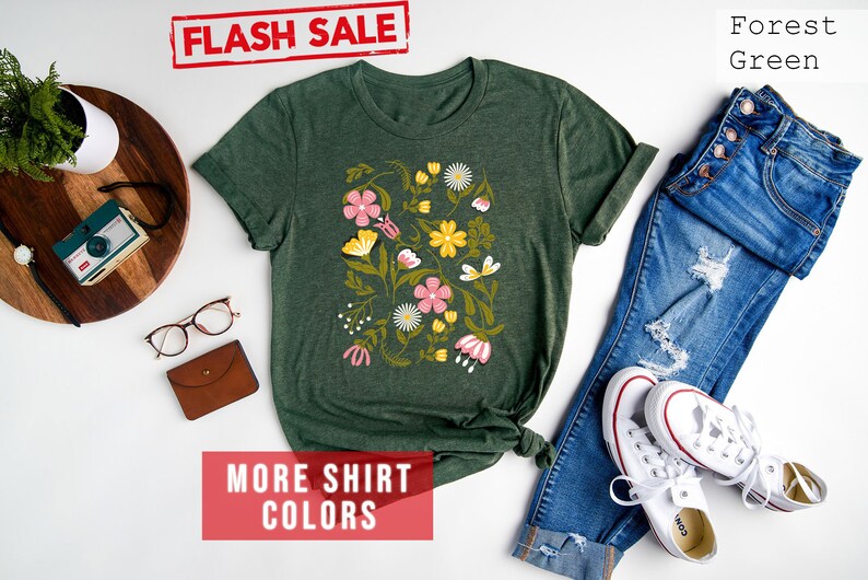 May include: Forest green t-shirt with a floral print featuring pink, yellow, and white flowers. The shirt is displayed with blue jeans and white sneakers. The text "FLASH SALE" and "MORE SHIRT COLORS" is visible in the image.