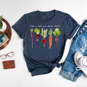 May include: Heather navy t-shirt with the text "Let's root for each other" and a watercolor illustration of various root vegetables. The shirt is paired with blue jeans and white sneakers.