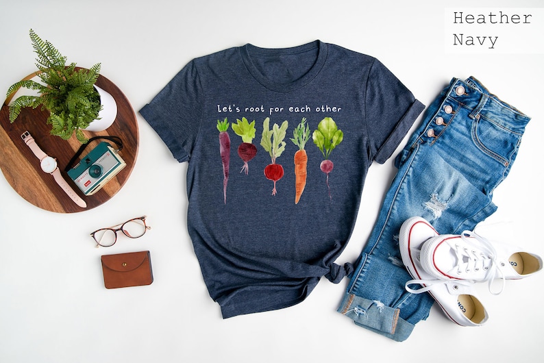 May include: Heather navy t-shirt with the text "Let's root for each other" and a watercolor illustration of various root vegetables. The shirt is paired with blue jeans and white sneakers.
