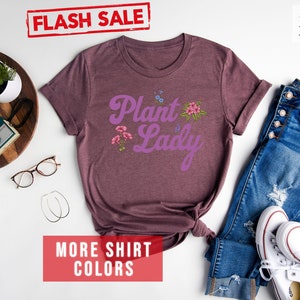 May include: A heather maroon t-shirt with the text "Plant Lady" in purple script with floral accents. The shirt is laid flat on a white surface with a pair of glasses, a wooden tray with a camera and a plant, and a pair of white sneakers and blue jeans.