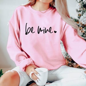 May include: Pink sweatshirt with the text "Be mine" in black script with a small heart.