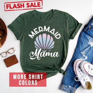 May include: Forest green t-shirt with the text "Mermaid Mama" in white with a pink and blue seashell graphic. The shirt is laid flat on a white surface with a pair of blue jeans, white sneakers, and other accessories.