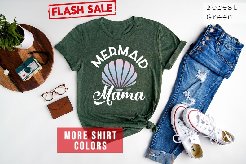 May include: Forest green t-shirt with the text "Mermaid Mama" in white with a pink and blue seashell graphic. The shirt is laid flat on a white surface with a pair of blue jeans, white sneakers, and other accessories.