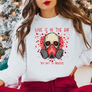 Pu&ograve; includere: Felpa bianca con un teschio rosso che indossa una maschera antigas e il testo "LOVE IS IN THE AIR TRY NOT TO BREATHE" circondato da cuori rossi e farfalle.