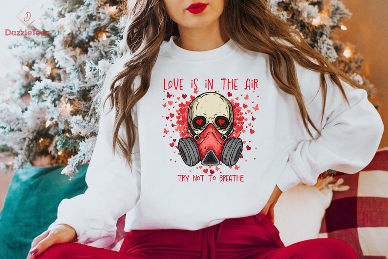 Pu&ograve; includere: Felpa bianca con un teschio rosso che indossa una maschera antigas e il testo "LOVE IS IN THE AIR TRY NOT TO BREATHE" circondato da cuori rossi e farfalle.