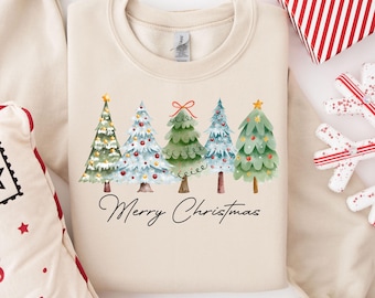 May include: A cream-colored sweatshirt featuring a watercolor design of five Christmas trees. Each tree is decorated with ornaments and topped with a star. The words "Merry Christmas" are written in a cursive font below the trees.