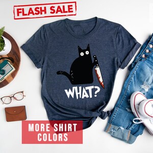 Op de afbeelding: Een heideblauw t-shirt met een zwarte kat die een bloedig mes vasthoudt en de tekst "WHAT?" erop gedrukt.