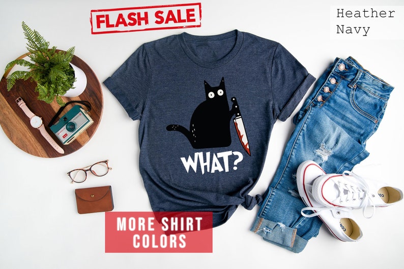 Op de afbeelding: Een heideblauw t-shirt met een zwarte kat die een bloedig mes vasthoudt en de tekst "WHAT?" erop gedrukt.