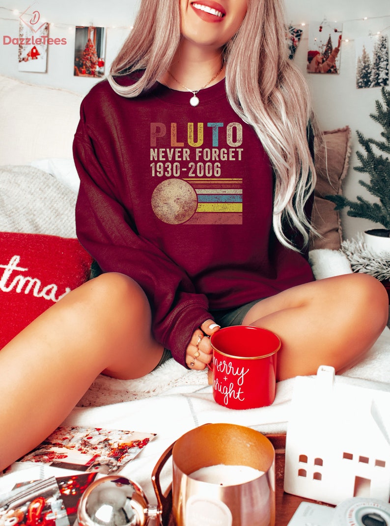 Op de afbeelding: Een bordeauxrood sweatshirt met een vintage-stijl grafische afbeelding met de tekst "Pluto Never Forget 1930-2006". De afbeelding toont een gestileerde afbeelding van Pluto met een regenboogkleurig streepontwerp.