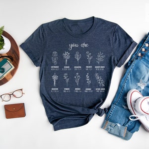 May include: A heather navy blue t-shirt with a white graphic design featuring flowers and the text "you are" with bible verses listed below each flower.
