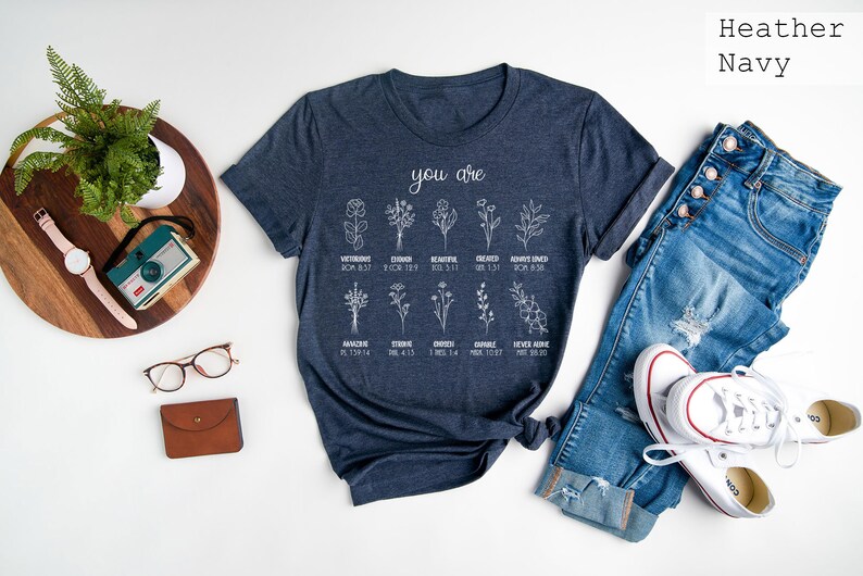 May include: A heather navy blue t-shirt with a white graphic design featuring flowers and the text "you are" with bible verses listed below each flower.
