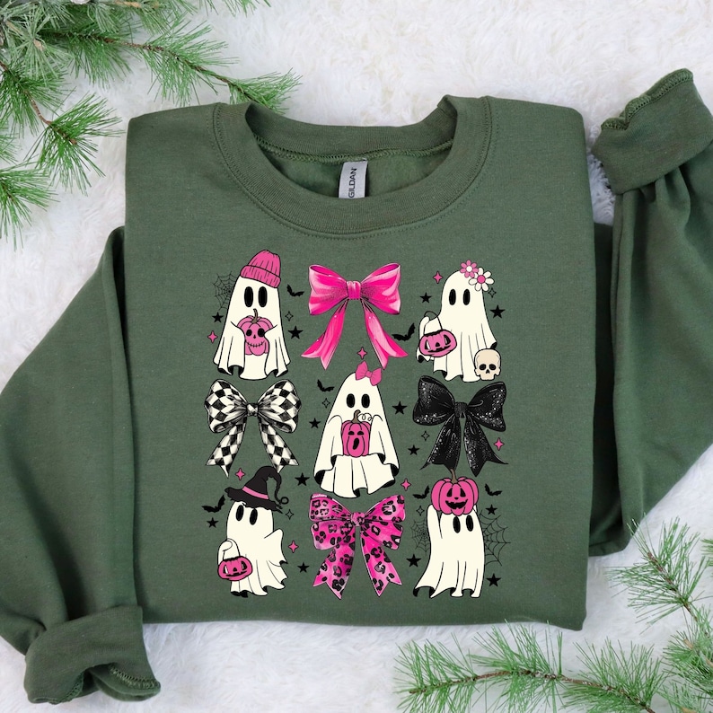 Puede incluir: Una sudadera verde oliva con un dise&ntilde;o de Halloween. El dise&ntilde;o incluye fantasmas de dibujos animados con lazos rosas, sombreros y bolsas de golosinas con forma de calabaza. Telara&ntilde;as negras y estrellas completan el look festivo.