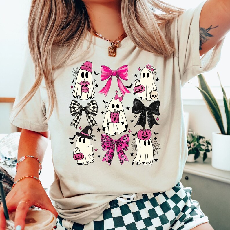 Puede incluir: Camiseta beige con un dise&ntilde;o tem&aacute;tico de Halloween. El gr&aacute;fico incluye varios personajes fantasma con acentos rosas, lazos y calabazas. El dise&ntilde;o tambi&eacute;n incorpora lazos a cuadros en blanco y negro, telara&ntilde;as y estrellas, creando una est&eacute;tica festiva y juguetona.
