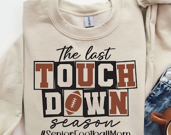 May include: Beige sweatshirt with the text "The last Touch Down season #SeniorFootballMom" in black and brown lettering. A football graphic is included in the text.