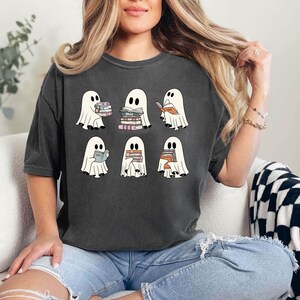 K&ouml;nnte beinhalten: Dunkelgraues T-Shirt mit sechs Cartoon-Geistern, die jeweils B&uuml;cher halten. Die Geister sind wei&szlig; mit schwarzen Augen und lesen oder halten B&uuml;cher mit farbigen Buchr&uuml;cken. Das T-Shirt ist ein l&auml;ssiger, &uuml;bergro&szlig;er Stil.