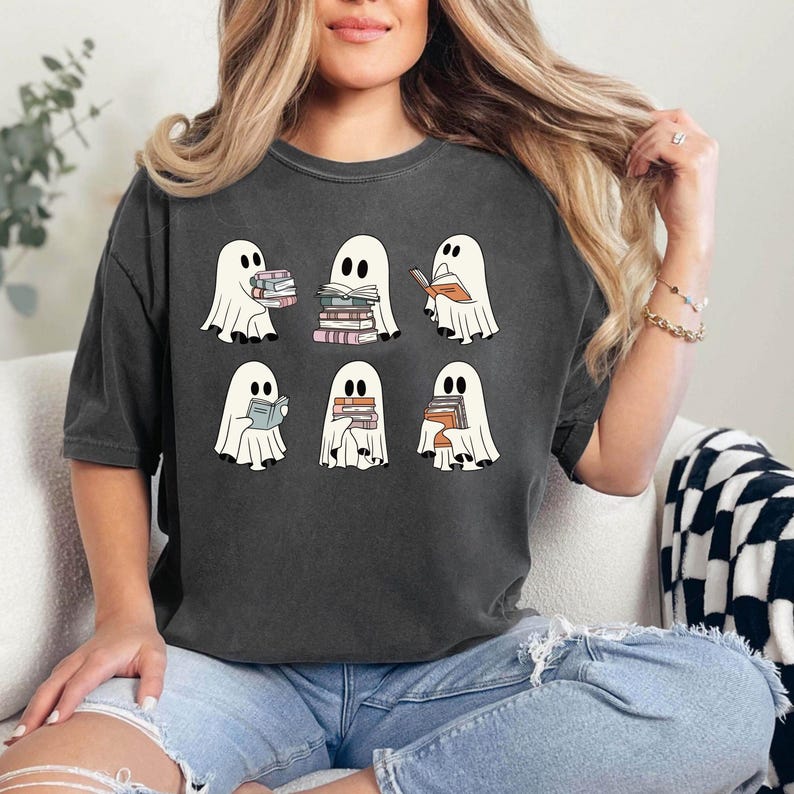 K&ouml;nnte beinhalten: Dunkelgraues T-Shirt mit sechs Cartoon-Geistern, die jeweils B&uuml;cher halten. Die Geister sind wei&szlig; mit schwarzen Augen und lesen oder halten B&uuml;cher mit farbigen Buchr&uuml;cken. Das T-Shirt ist ein l&auml;ssiger, &uuml;bergro&szlig;er Stil.