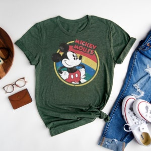 Puede incluir: Una camiseta verde con un gr&aacute;fico de estilo vintage de Mickey Mouse. El gr&aacute;fico presenta a Mickey Mouse con un atuendo rojo y amarillo con un arco&iacute;ris detr&aacute;s de &eacute;l. El texto "Mickey Mouse" est&aacute; escrito en una fuente retro sobre Mickey Mouse.