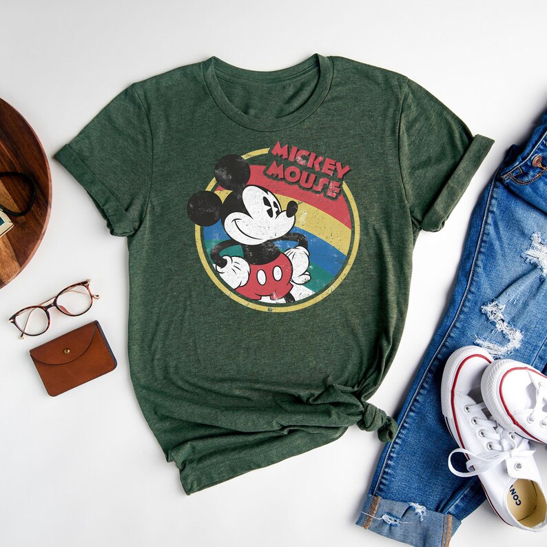 Puede incluir: Una camiseta verde con un gr&aacute;fico de estilo vintage de Mickey Mouse. El gr&aacute;fico presenta a Mickey Mouse con un atuendo rojo y amarillo con un arco&iacute;ris detr&aacute;s de &eacute;l. El texto "Mickey Mouse" est&aacute; escrito en una fuente retro sobre Mickey Mouse.