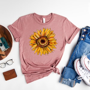 May include: A heather mauve t-shirt with a yellow sunflower graphic. The shirt is laid flat on a white surface with a pair of jeans, a pair of white sneakers, a wooden tray with a plant and a camera, and a pair of glasses.