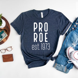 Op de afbeelding: Een marineblauw t-shirt met witte tekst die "PRO ROE est. 1973" luidt. Het shirt ligt plat op een wit oppervlak met een spijkerbroek, een horloge, een camera en een bril.