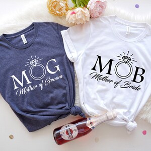 May include: Two white t-shirts with black text and a diamond ring graphic. The text on the left shirt reads "MOG Mother of Groom". The text on the right shirt reads "MOB Mother of Bride".