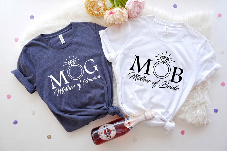 May include: Two white t-shirts with black text and a diamond ring graphic. The text on the left shirt reads "MOG Mother of Groom". The text on the right shirt reads "MOB Mother of Bride".