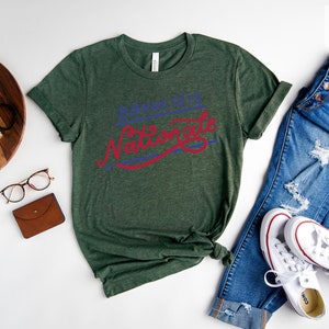 May include: A forest green short-sleeve t-shirt featuring the text Bonne F&ecirc;te Nationale in blue and red script lettering. The shirt is displayed alongside blue denim jeans, white sneakers, a small camera, glasses, and a brown wallet.