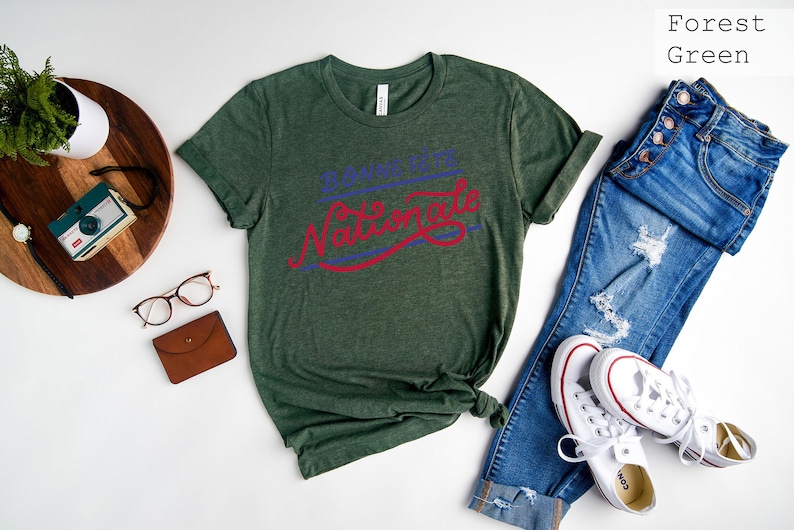 May include: A forest green short-sleeve t-shirt featuring the text Bonne F&ecirc;te Nationale in blue and red script lettering. The shirt is displayed alongside blue denim jeans, white sneakers, a small camera, glasses, and a brown wallet.