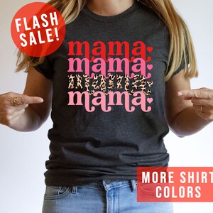 Op de afbeelding: Een donkergrijs T-shirt met de tekst "mama mama mama" in roze en rode letters met een luipaardprint en harten. De tekst is in een retro-lettertype.