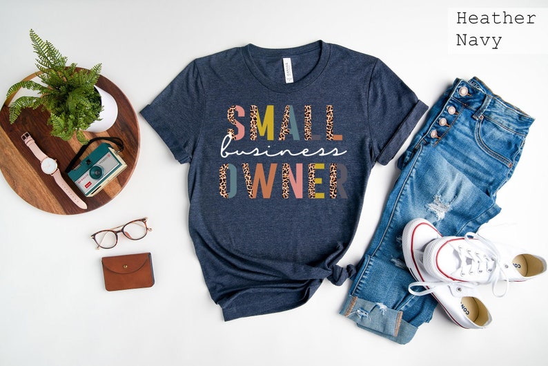 Pode incluir: Uma camiseta azul marinho heather com o texto "SMALL business OWNER" em letras coloridas com um padr&atilde;o de leopardo.