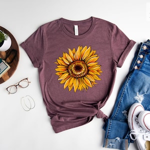 May include: A heather maroon t-shirt with a sunflower graphic. The shirt is laid flat on a white surface with a pair of jeans, a pair of white sneakers, and a small potted plant.