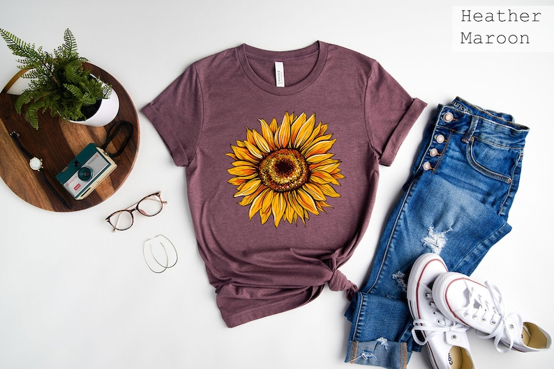 May include: A heather maroon t-shirt with a sunflower graphic. The shirt is laid flat on a white surface with a pair of jeans, a pair of white sneakers, and a small potted plant.