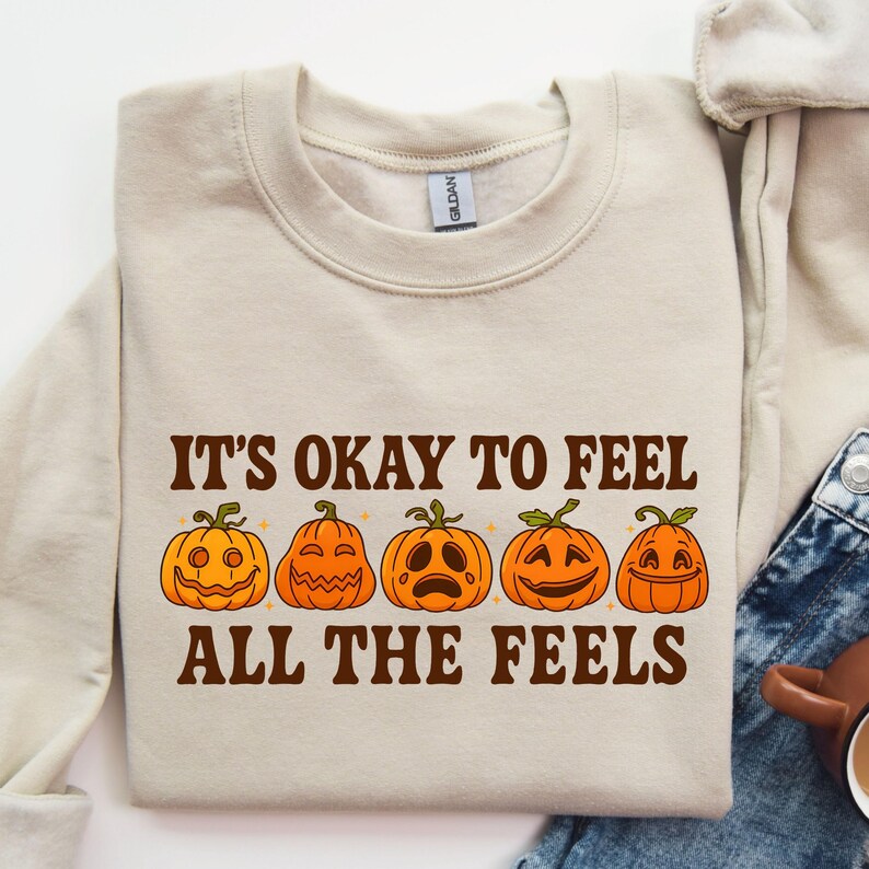 May include: Beige sweatshirt with the text "IT'S OKAY TO FEEL ALL THE FEELS" in brown. Below the text are five orange jack-o'-lanterns with different facial expressions. The sweatshirt is made of a soft material.