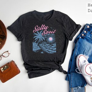 May include: A dark grey t-shirt with a graphic of a palm tree, waves, and a sun. The text "Salty Soul" is printed in pink on the shirt.