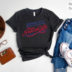 May include: A heather dark grey short-sleeve t-shirt featuring the text "BONNE F&Ecirc;TE" in blue and "Nationale" in red script. The shirt is displayed alongside blue jeans, white sneakers, a brown wallet, glasses, a vintage camera, and a succulent.