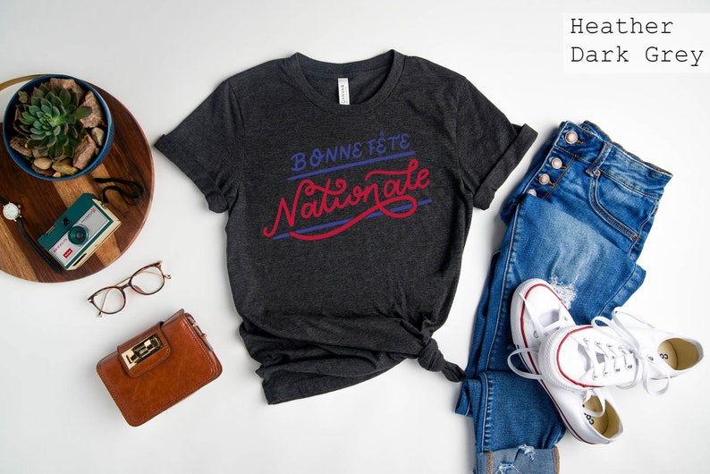 May include: A heather dark grey short-sleeve t-shirt featuring the text "BONNE F&Ecirc;TE" in blue and "Nationale" in red script. The shirt is displayed alongside blue jeans, white sneakers, a brown wallet, glasses, a vintage camera, and a succulent.